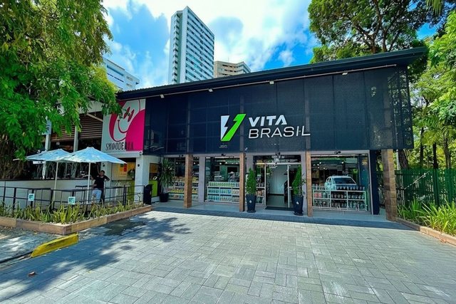 Fachada de uma loja VitaBrasil sob árvores, com calçada larga e vitrines abertas.
