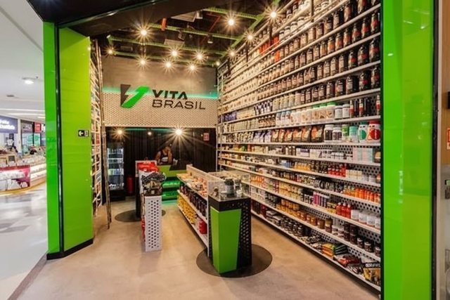 Interior de uma loja VitaBrasil: corredor com prateleiras altas dos dois lados, cheias de potes e caixas de suplementos de marcas diferentes.
